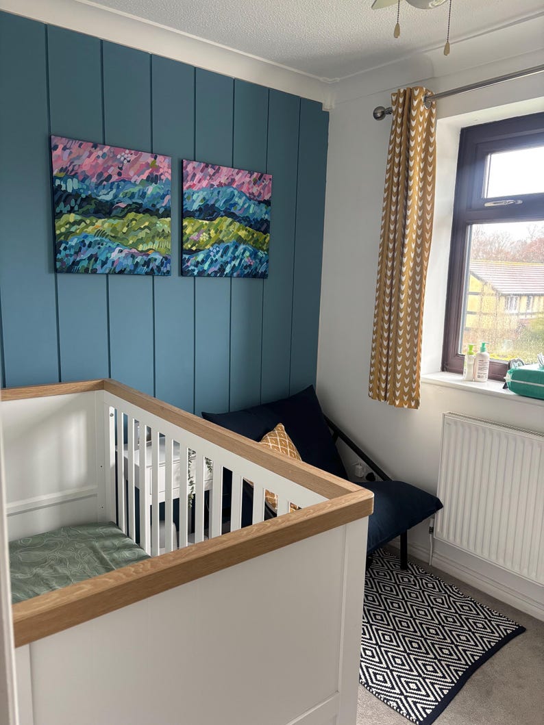 Puede incluir: Un cuarto de beb&eacute; con una cuna blanca con detalles de madera, una pared decorativa azul y dos pinturas abstractas de paisajes. Una silla azul marino con un coj&iacute;n estampado se encuentra junto a una ventana con cortinas amarillas. Una alfombra geom&eacute;trica en blanco y negro est&aacute; en el suelo.