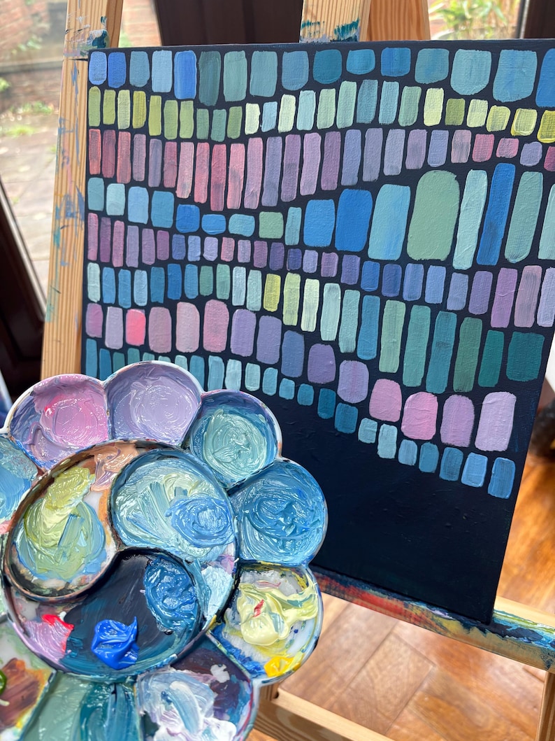 Op de afbeelding: Abstract schilderij op zwart canvas met verticale rechthoeken in blauw, groen, roze en paars. Een bloemvormig kunstenaarspalet met diverse verfkleuren staat voor het canvas.