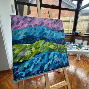 Puede incluir: Una pintura abstracta de paisaje sobre lienzo, exhibida en un caballete de madera. La obra presenta capas de color, incluyendo azules, verdes y morados, creando un efecto texturizado. La pintura est&aacute; en una habitaci&oacute;n bien iluminada con vistas al exterior.
