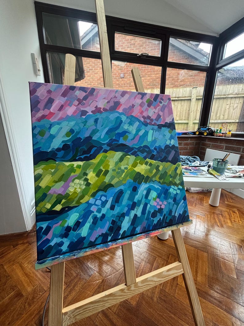 Puede incluir: Una pintura abstracta de paisaje sobre lienzo, exhibida en un caballete de madera. La obra presenta capas de color, incluyendo azules, verdes y morados, creando un efecto texturizado. La pintura est&aacute; en una habitaci&oacute;n bien iluminada con vistas al exterior.