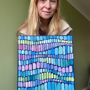 Op de afbeelding: Vierkant abstract schilderij met een donkerblauwe rand. Het kunstwerk toont een moza&iuml;ek van kleurrijke, verticale rechthoeken in tinten blauw, groen, roze en paars. Het schilderij wordt vastgehouden door een persoon met blond haar.