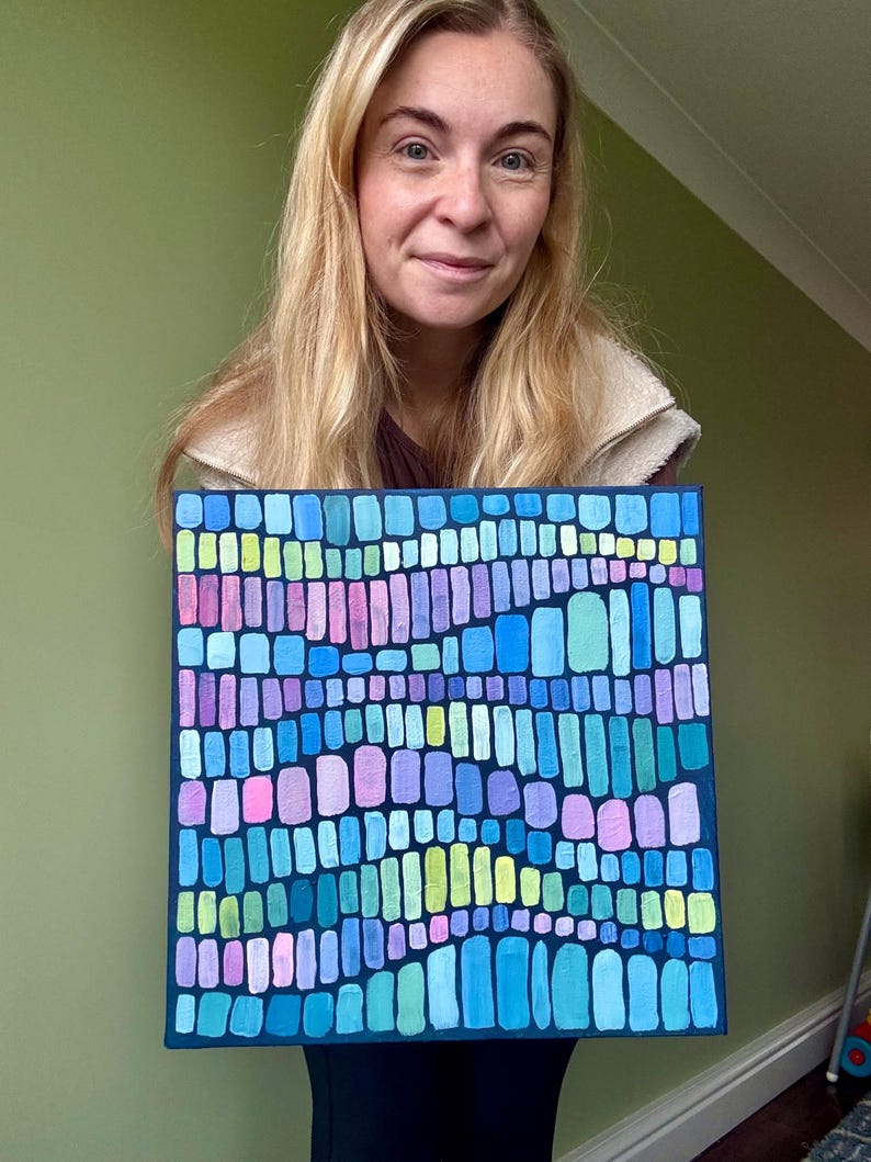 Op de afbeelding: Vierkant abstract schilderij met een donkerblauwe rand. Het kunstwerk toont een moza&iuml;ek van kleurrijke, verticale rechthoeken in tinten blauw, groen, roze en paars. Het schilderij wordt vastgehouden door een persoon met blond haar.