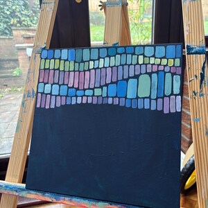 Op de afbeelding: Een abstract schilderij op canvas, tentoongesteld op een houten ezel. Het kunstwerk heeft een donker marineblauwe achtergrond met een bovenste gedeelte van kleurrijke rechthoekige vormen in tinten blauw, groen en paars. Het schilderij is een modern kunstwerk.