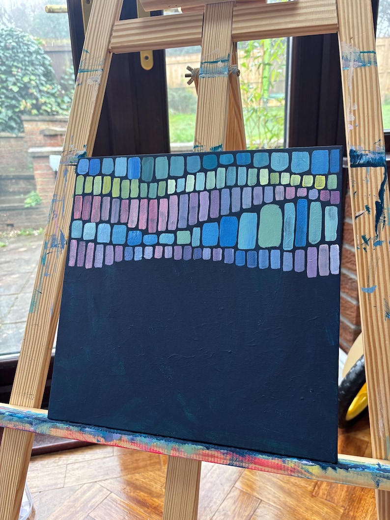 Op de afbeelding: Een abstract schilderij op canvas, tentoongesteld op een houten ezel. Het kunstwerk heeft een donker marineblauwe achtergrond met een bovenste gedeelte van kleurrijke rechthoekige vormen in tinten blauw, groen en paars. Het schilderij is een modern kunstwerk.