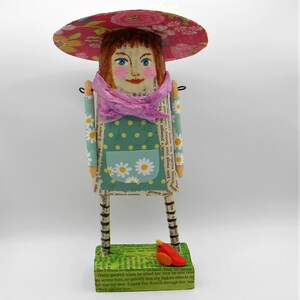 May include: A whimsical paper doll figurine with a pink and floral hat, a pink scarf, and a green and white polka dot dress. The doll is standing on a green base with a small red bird figurine.