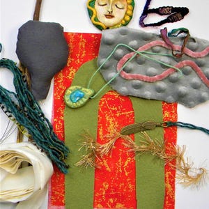 May include: A collection of craft supplies including a sculpted face pendant, a grey textured fabric piece, a red and gold patterned fabric, green felt shapes, a blue stone pendant, a metal feather charm, yarn, ribbon, and a wooden stick.