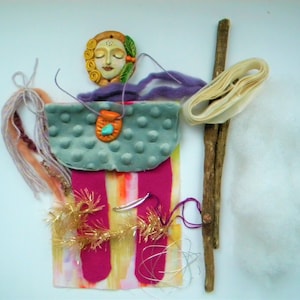 May include: A craft kit featuring a sculpted clay face with yellow hair and green leaves, a teal dimpled fabric piece, two magenta felt strips, a wooden stick, cream ribbon, white fiber fill, purple yarn, gold tinsel, and a small clay pendant.