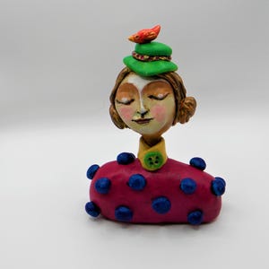 May include: A handmade ceramic figurine of a woman with closed eyes wearing a green hat with a red bird on top. She is wearing a pink dress with blue polka dots and a yellow collar with a green button.