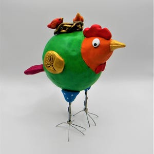 May include: A green, orange, and red clay chicken with a nest and two small birds on its head. The chicken has blue and white polka dot legs and wire feet.