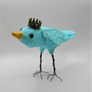 May include: A whimsical, light blue bird sculpture with a textured surface. The bird has a gold-colored beak and a small, ornate gold crown. Its legs are thin, dark wire, and it stands on a white surface. A unique decorative item.