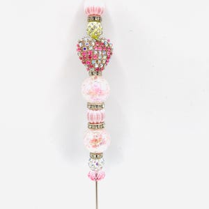 May include: A decorative pen featuring a vertical arrangement of beads in various shades of pink, white, and lime green. The design includes a central heart-shaped bead covered in iridescent crystals, interspersed with silver-toned spacers and clear beads.