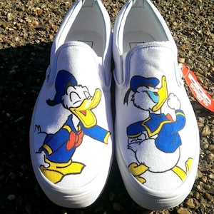 May include: A pair of white canvas slip-on sneakers featuring a graphic illustration of Donald Duck on the front of each shoe. The character is depicted in a blue sailor outfit with a yellow beak and feet. A red tag is attached to the side.