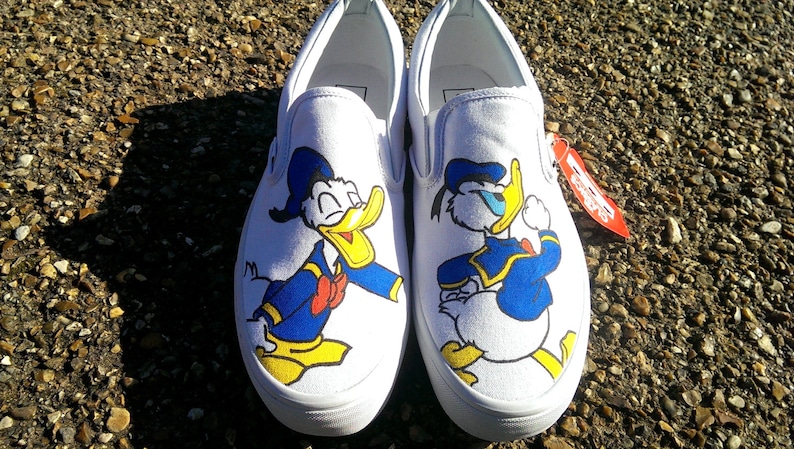May include: A pair of white canvas slip-on sneakers featuring a graphic illustration of Donald Duck on the front of each shoe. The character is depicted in a blue sailor outfit with a yellow beak and feet. A red tag is attached to the side.