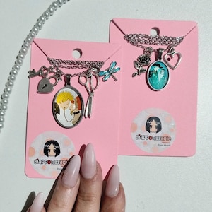 Pode incluir: Dois colares em cartões rosa. Um apresenta um pingente com um casal, um cadeado, uma tesoura e um pingente de libélula. O outro tem uma rosa, um coração e um pingente oval com um design turquesa. Cada cartão tem um adesivo com uma menina de desenho animado e o texto "giapponeserie".