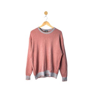 Può includere: Un maglione a righe corallo e grigio è appeso a una gruccia di legno. Il maglione a maniche lunghe ha una trama a coste con girocollo, polsini e orlo grigi. Il maglione è esposto su uno sfondo bianco.