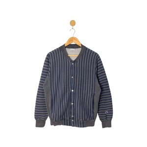 Puede incluir: Chaqueta abotonada a rayas grises y azul marino. La chaqueta tiene cuello, puños y dobladillo de canalé. El logo de Champion es visible en la manga inferior derecha. La chaqueta está colgada en una percha de madera.