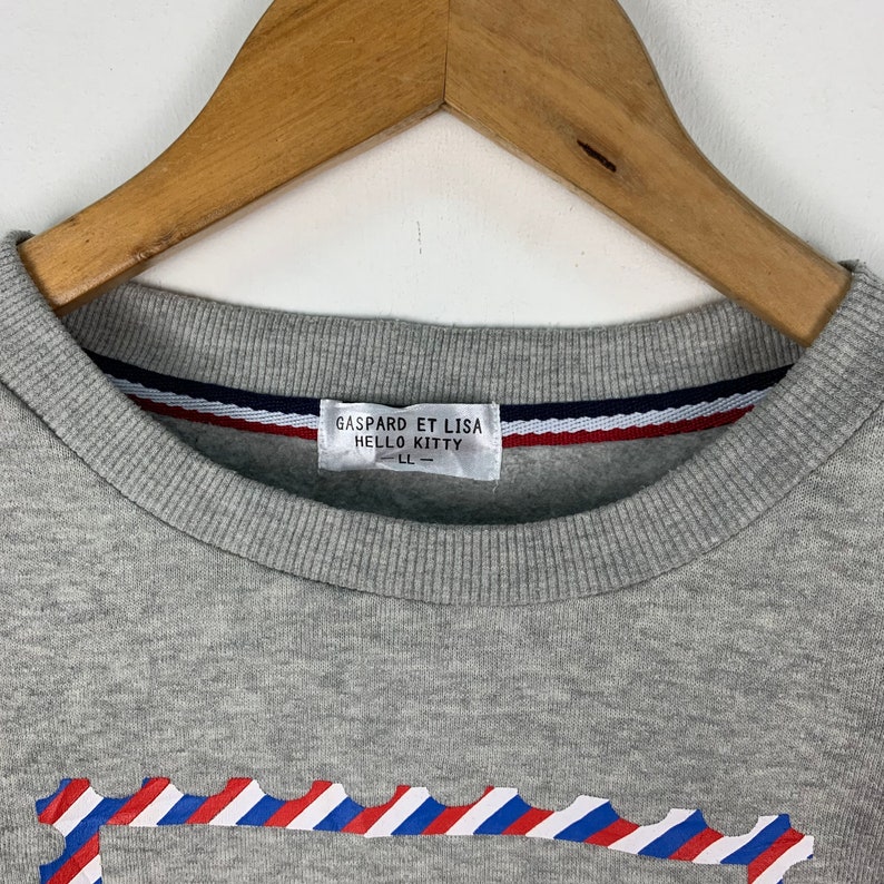 Puede incluir: Sudadera gris con un dise&ntilde;o a rayas rojo, blanco y azul. La sudadera tiene una etiqueta que dice "Gaspard et Lisa Hello Kitty - LL - "