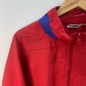 Op de afbeelding: Rode windbreaker-jas met een blauw mesh-paneel op de capuchon en een ritssluiting. De jas heeft een label met de tekst "1516V".