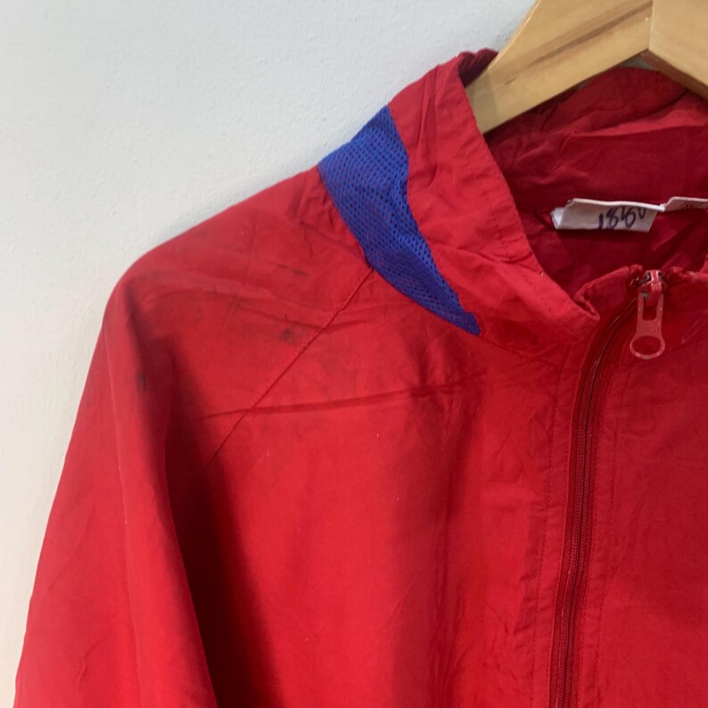 Op de afbeelding: Rode windbreaker-jas met een blauw mesh-paneel op de capuchon en een ritssluiting. De jas heeft een label met de tekst "1516V".