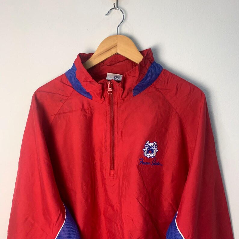 Op de afbeelding: Rode windbreaker-jas met blauwe accenten en een geborduurd bulldog-logo in wit en blauw met de tekst "Fremont State".