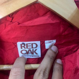 Op de afbeelding: Red Oak Sportswear-label op een rood jack met een maatlabel waarop XL staat.