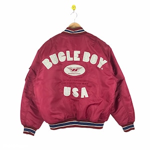 May include: A red bomber jacket with a white "Buckle Boy" logo on the back. The logo includes the text "International Industries" and "Style Favorite The American Way" in a smaller font. The jacket has a blue and white striped collar and cuffs.