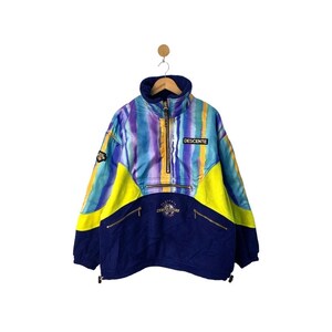 Könnte beinhalten: Eine blaue, gelbe und violette Skijacke mit einem Batik-Muster. Die Jacke hat einen Reißverschluss und einen hohen Kragen. Der Markenname "DESCENTE" ist auf der Vorderseite der Jacke aufgedruckt.