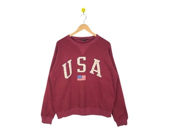 Può includere: Una felpa color granata con una grande scritta ricamata "USA" in bianco e una piccola bandiera americana sul davanti.