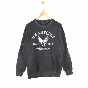 May include: A charcoal grey sweatshirt featuring the white text "U.S. AIR FORCE" and a graphic emblem. Additional text includes "R.A.F. 18th" and "Lakenheath A.F. Base Royal Air Force". The garment hangs on a wooden hanger.