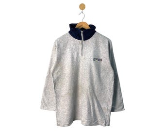 Puede incluir: Un jersey de polar gris con cierre de cremallera a la mitad y cuello azul. El jersey tiene la palabra "Remarkable" impresa en la parte delantera en letras de colores.