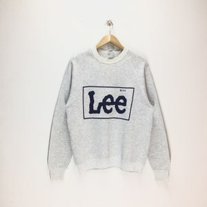 May include: A light gray sweatshirt with the brand name "Lee" in a navy blue font inside a navy blue rectangle. The sweatshirt has a crew neck and long sleeves. The Lee logo is centered on the front.