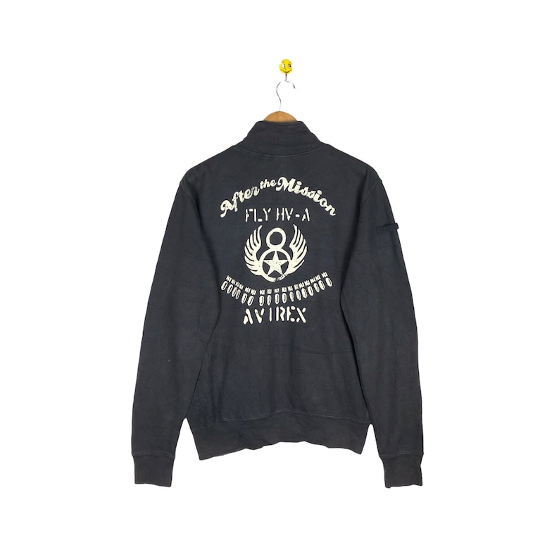 May include: A black zippered sweatshirt with a white graphic on the back that says "After the Mission Fly HV-A" with a star and wings. The graphic also includes the word "AVIREX" in white.