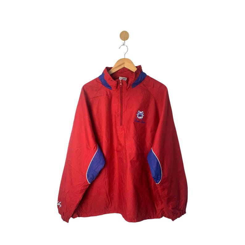 Op de afbeelding: Rode windbreaker-jas met blauwe accenten en een klein geborduurd logo op de voorkant. De jas heeft een ritssluiting en een capuchon met trekkoord.