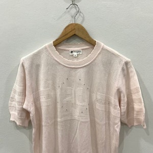 May include: A pale pink short-sleeved sweater with a white embroidered design on the front. The design features a heart and the word "Couronne" in a script font.