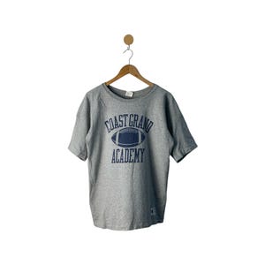 Puede incluir: Camiseta gris de manga corta con un gráfico azul marino de un balón de fútbol y el texto "Coast Grand Academy".