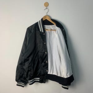 May include: Black satin baseball jacket with white stripes on the collar and cuffs. The jacket has a snap closure and multiple pockets.
