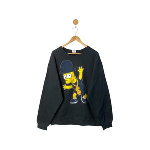 Könnte beinhalten: Schwarzes Sweatshirt mit einer Grafik von Bart Simpson in einer Hip-Hop-Pose. Bart trägt einen dunkelblauen Hut, eine goldene Kette und dunkelblaue Shorts. Das Sweatshirt hängt an einem Holzbügel vor weißem Hintergrund.