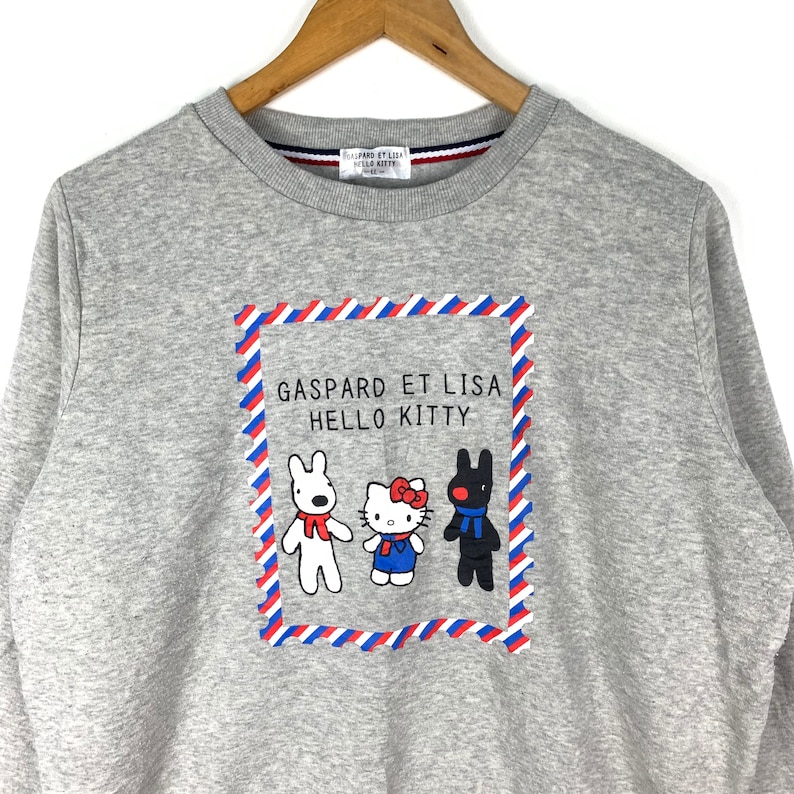 Puede incluir: Sudadera gris con un estampado gr&aacute;fico de un sello postal rojo, blanco y azul con los personajes Gaspard et Lisa y Hello Kitty.