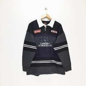 May include: A long-sleeved rugby shirt in black, gray, and navy blue, with a white collar. The shirt features the "Canterbury of New Zealand" logo and "CCC SPORT" and "Steinlager" patches.