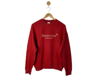 Puede incluir: Una sudadera roja de Benetton con el nombre de la marca y "United Colors" bordado en un color más claro. La sudadera de cuello redondo está colgada de una percha de madera sobre un fondo blanco. La sudadera de estilo vintage es de color sólido.