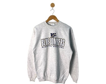 May include: A heather gray sweatshirt with the text "BLUE BOMBERS" in black and white, with a blue and gold logo above the text. The text "WINNIPEG, MANITOBA" is below the main text. The sweatshirt is hanging on a wooden hanger.