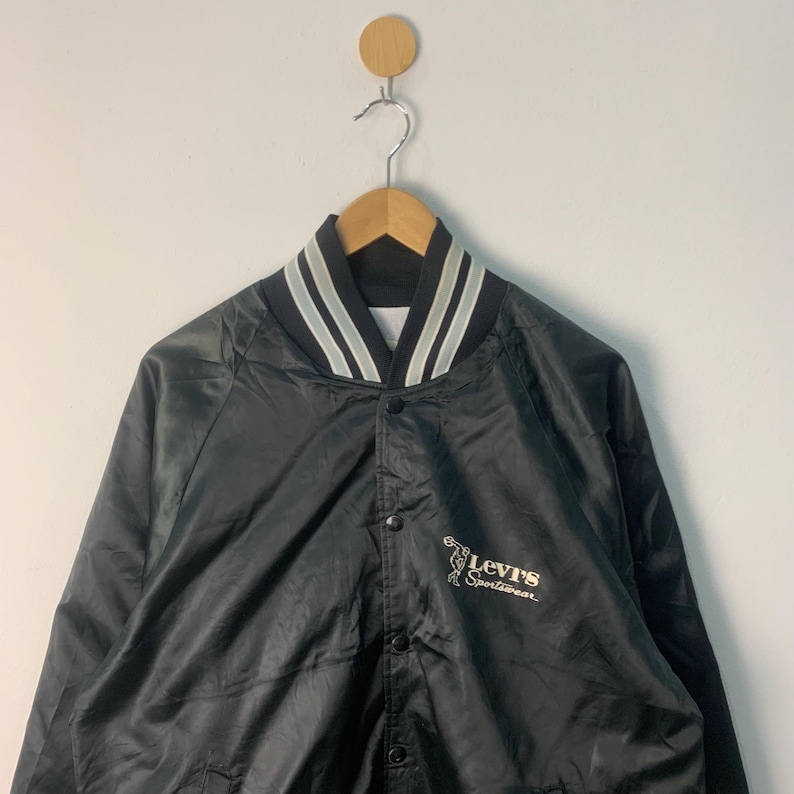 May include: A black satin baseball jacket with white stripes on the collar and a Levi's sportswear logo on the right side.