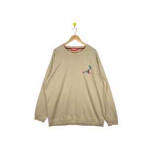 May include: A beige long-sleeve sweatshirt with a small embroidered graphic on the left chest. The graphic features a cartoon character with a red hat and a blue skateboard.