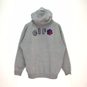 Op de afbeelding: Een grijze hoodie met het woord "elf" in marineblauwe letters en een rood-blauw geometrisch ontwerp. De hoodie heeft een capuchon en lange mouwen.