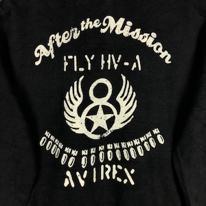 May include: Black t-shirt with white text that reads "After the Mission FLY HV-A" and a white graphic of a star with wings. The text "AVIREX" is printed below the graphic.