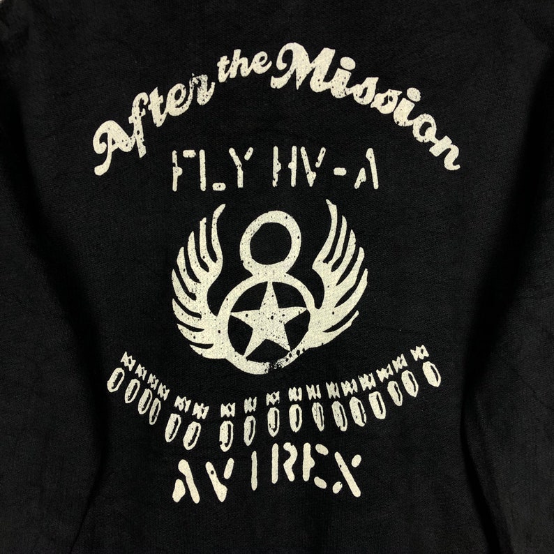 May include: Black t-shirt with white text that reads "After the Mission FLY HV-A" and a white graphic of a star with wings. The text "AVIREX" is printed below the graphic.