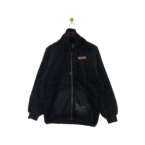 May include: Black fleece jacket with a zipper closure and a brown patch with the word "Avex" on the left chest. The jacket has a black pocket on the lower left side with the text "Red Hats" printed on it.