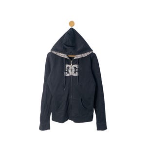 May include: Black zip-up hoodie with a hood and front pockets. The hoodie features a white and gray patterned design along the hood's edge and a white and gray logo on the front. The hoodie is hanging on a wooden peg.