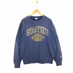 Puede incluir: Una sudadera azul Champion con un gráfico dorado y blanco de un balón de fútbol y el texto "GUSTIES DEFENSE".