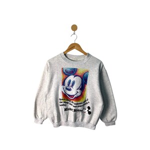 Peut inclure: Un sweat-shirt gris avec un motif coloré du visage de Mickey Mouse. Le texte "Since 1928, the beloved idol of the world has been everybody's leader, Mickey Mouse" est imprimé sous le motif.